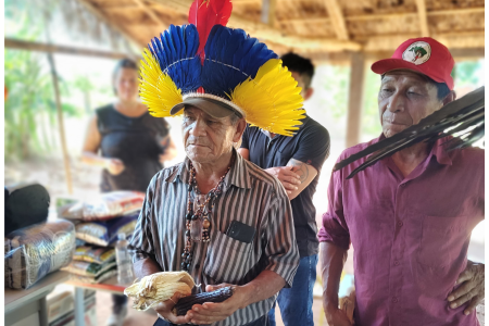 Dois homens indígenas estão em um espaço coberto. Um deles usa um cocar de penas azuis, amarelas e vermelhas e segura espigas de milho claras e escuras; o outro veste camisa roxa e boné vermelho. Ao fundo, há outras pessoas e sacos de produtos empilhados.