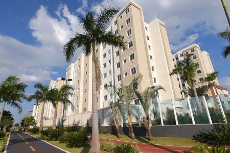 Vista externa de um condomínio da MRV, no primeiro plano tem umas palmeiras e uma calçada com grama verde e um passeio laranja, ao fundo prédios de nove andares nas cores bege e branca