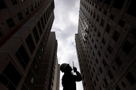 Mulher engenheira segura rádio de comunicação e fala com aparelho, ela está entre dois prédios em construção, a foto está levemente contra luz