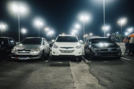 Fotografia mostra um estacionamento à noite com três carros emparelhados. Estacionados com a frente para a fotos, o carro da esquerda é cinza, o do meio é branco e o da direita é grafite. No último carro à direita algumas pessoas estão próximas. Ao fundo, luzes de postes acesas.