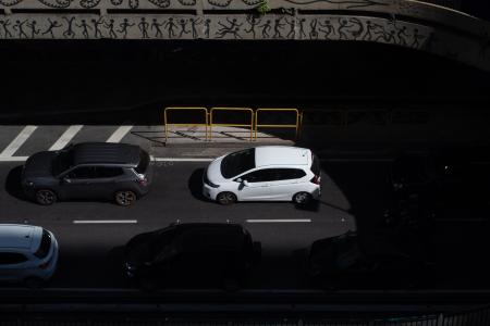 Imagem mostra cenário urbano com viaduto. Na parte de baixo, carros passam na pista, alguns deles estão encobertos pela sombra do viaduto, um carro cinza e um branco se destacam no centro iluminados pelo sol.
