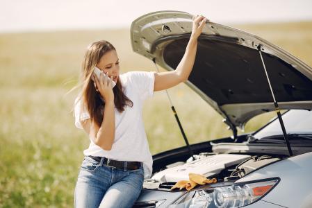 Imagem mostra uma mulher jovem na estrada com o capô do carro levantado. Ela está de pé, encostada na lataria e com a mão esquerda apoiada no capô aberto enquanto observa a parte interna do carro. Com a mão direita segura o celular no ouvido. Ela tem cabelos lisos castanhos soltos, veste uma blusa branca e está de calça jean com cinto. O fundo da imagem mostra uma paisagem com gramado verde.