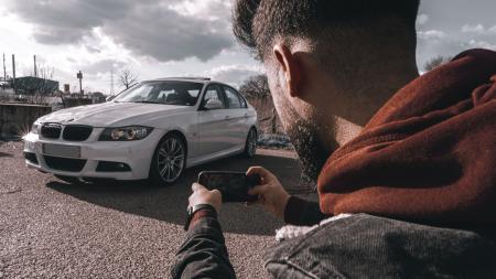 Imagem mostra um homem de costas, ela veste roupa de frio com jaqueta jeans. Possui cabelo baixo e barba, seguro o celular com as duas mãos e fotografa um carro branco em uma paisagem que mostra nuvens ao fundo.