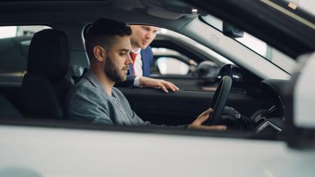Fotografia mostra um homem branco de perfil, sentado à frente da direção de um carro branco. Do lado de fora, homem branco de terno parece dar instruções