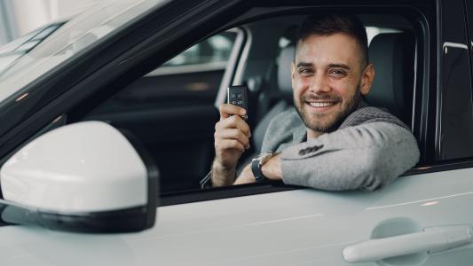 Imagem mostra um carro branco e dentro dele um homem de cabelos curtos e barba que veste um paletó. Ele segura a chave do carro com a mão direita e sorri para a câmera.Imagem mostra um carro branco e dentro dele um homem de cabelos curtos e barba que veste um paletó. Ele segura a chave do carro com a mão direita e sorri para a câmera.