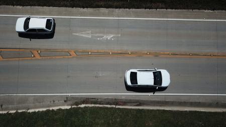Imagem mostra a estrada de cima. Dois carros brancos aparecem, cada um em uma pista, seguindo direções opostas.
