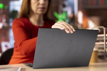 Mulher com blusa vermelha utiliza um notebook em um ambiente interno, com a mão apoiada na tela do computador e o fundo desfocado.