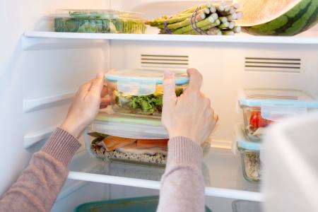 Pessoa organiza recipientes com refeições prontas, saladas e outros alimentos em prateleiras da geladeira, destacando o armazenamento adequado e o planejamento das refeições.