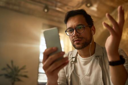 Homem jovem faz uma chamada de vídeo pelo celular, usando óculos de grau e fones de ouvido, enquanto gesticula com uma das mãos em um ambiente interno.